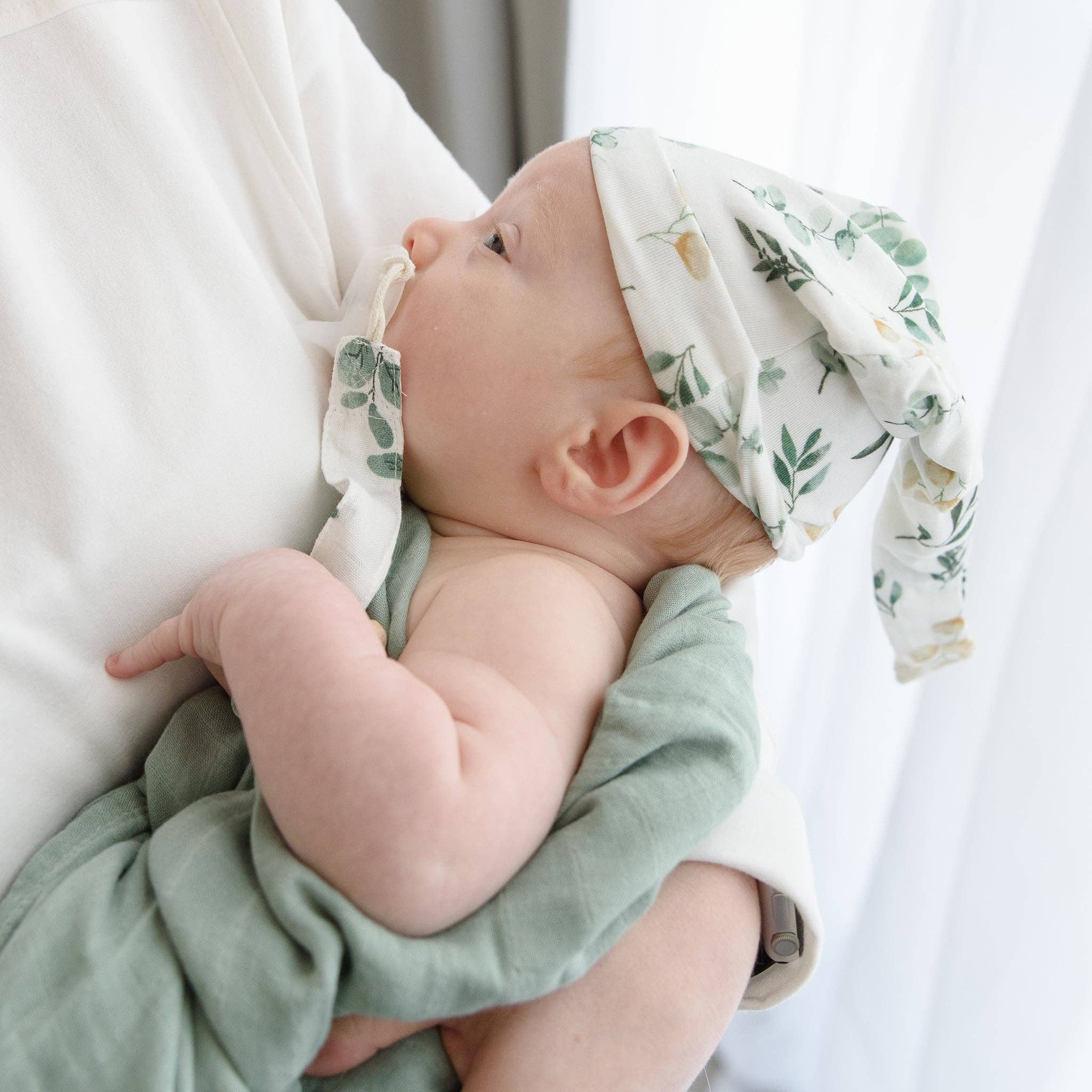 Baby wrapped in green bamboo cotton blanket with eucalyptus design, wearing matching hat, perfect for neutral baby gift set.