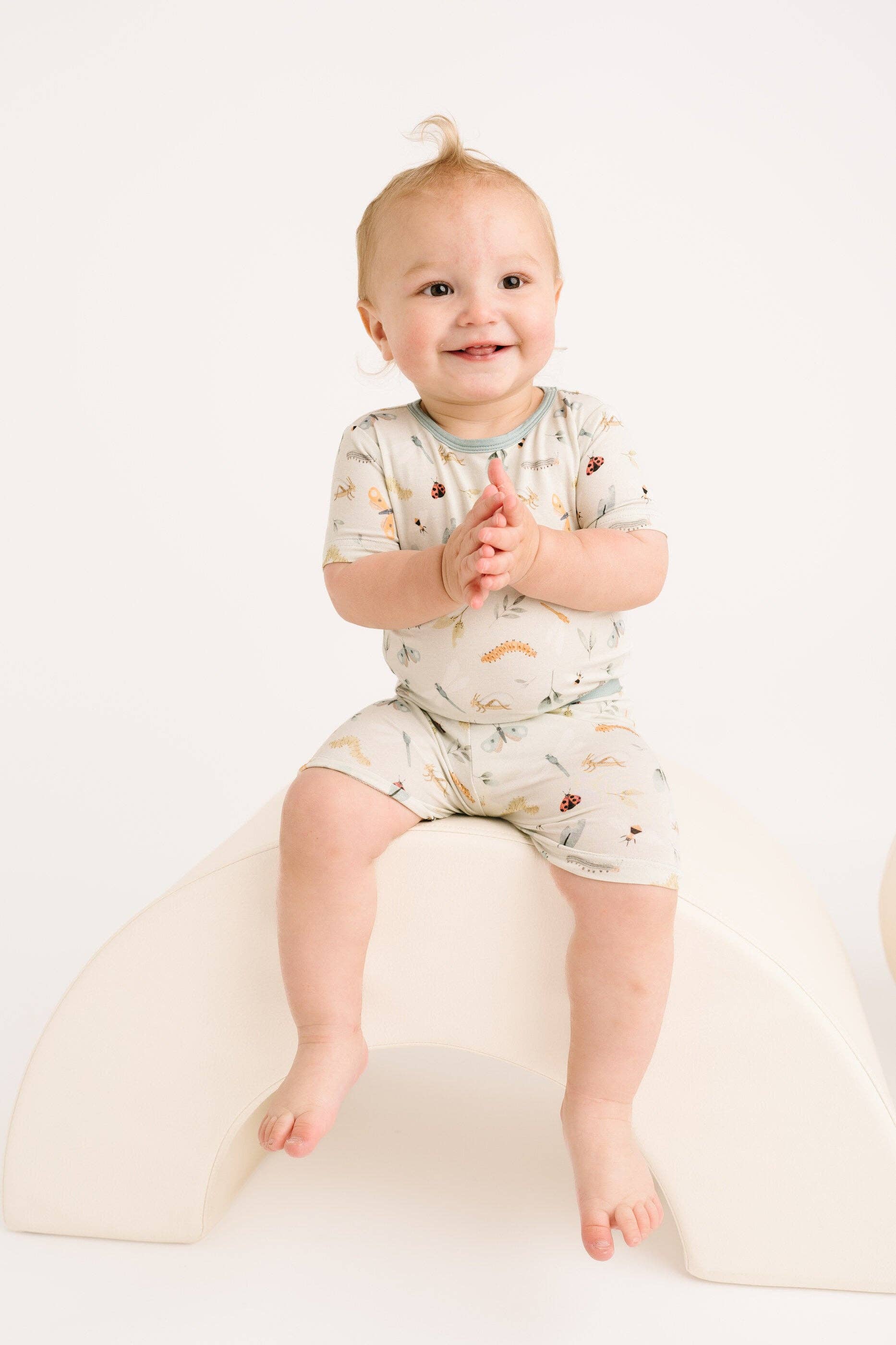 Smiling toddler in Bug Buddies Bamboo Shorts Set from Little One Shop, 12-18M, sitting on cream chair.