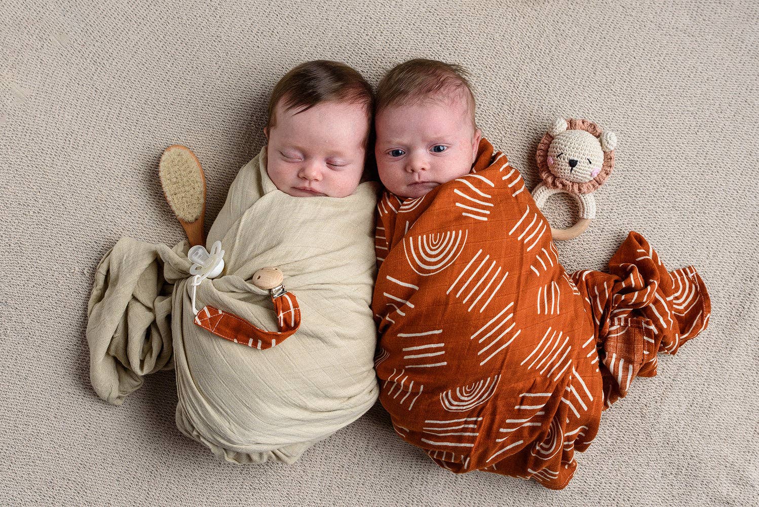 Two adorable newborns wrapped in Coco Stripes Terra swaddles, featuring boho design and plush toys. Perfect for a deluxe baby gift set.