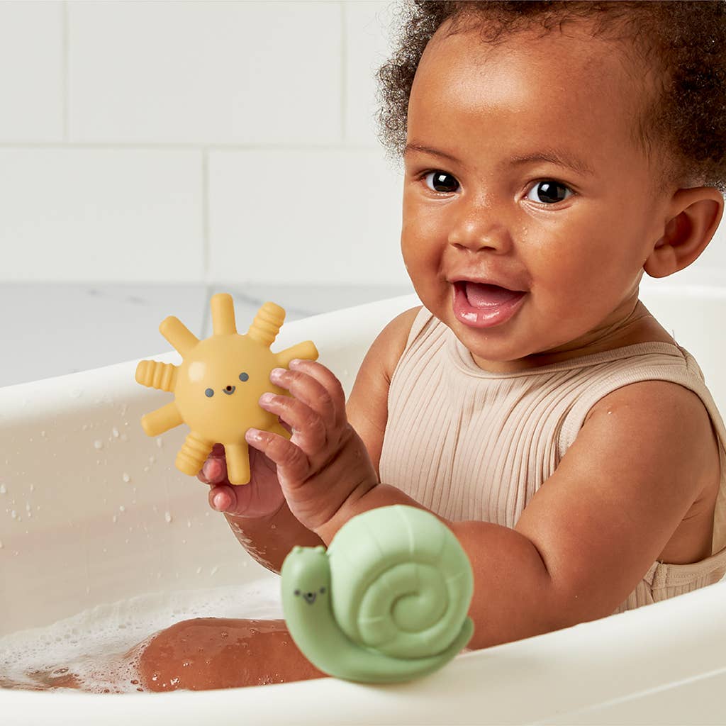 Happy baby in bath holding yellow sun squirt toy and green snail, enjoying playtime with Itzy Squirt™ bath toys.