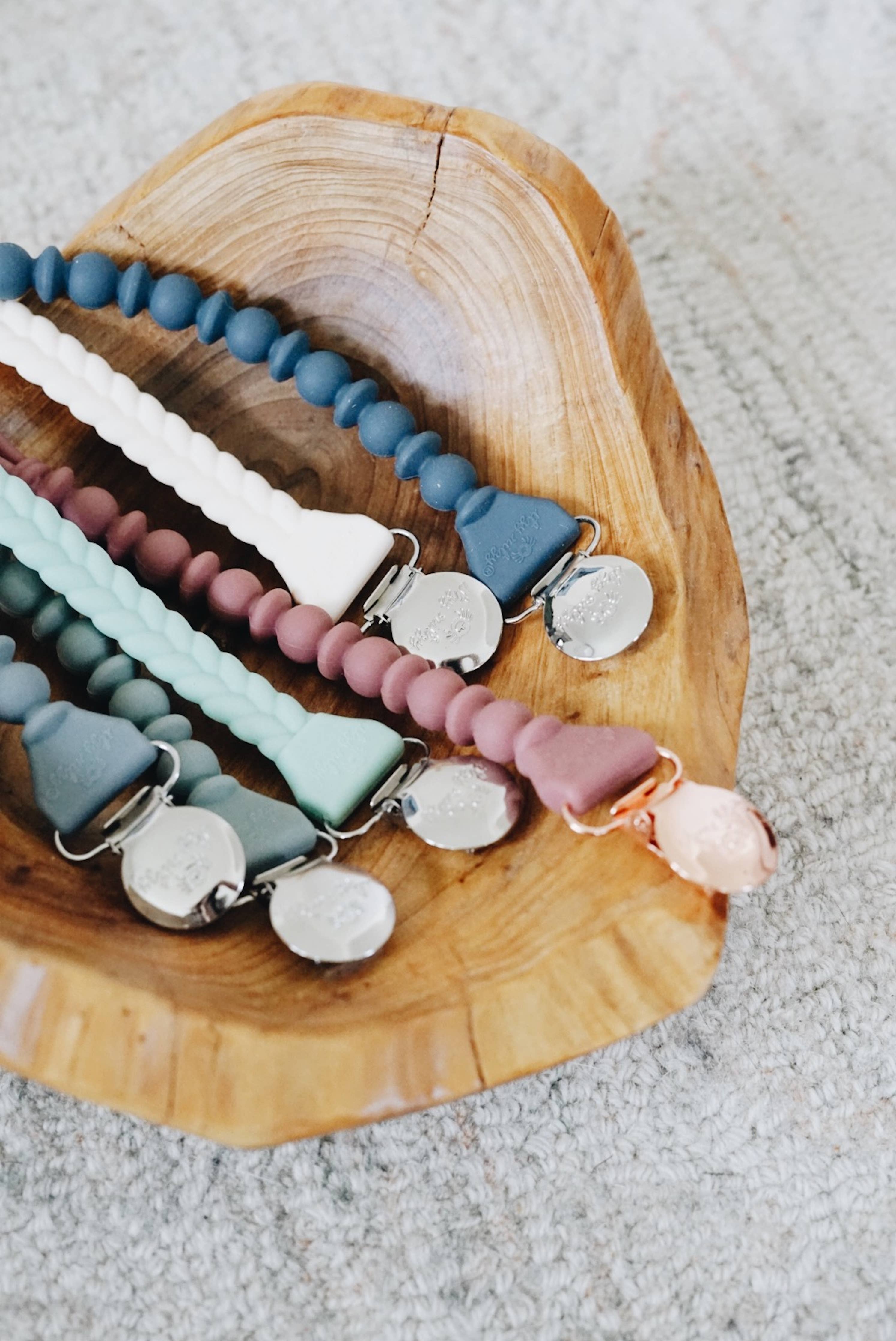 Colorful Sweetie Strap™ silicone pacifier clips displayed in a wooden bowl, showcasing their stylish braided and beaded design.