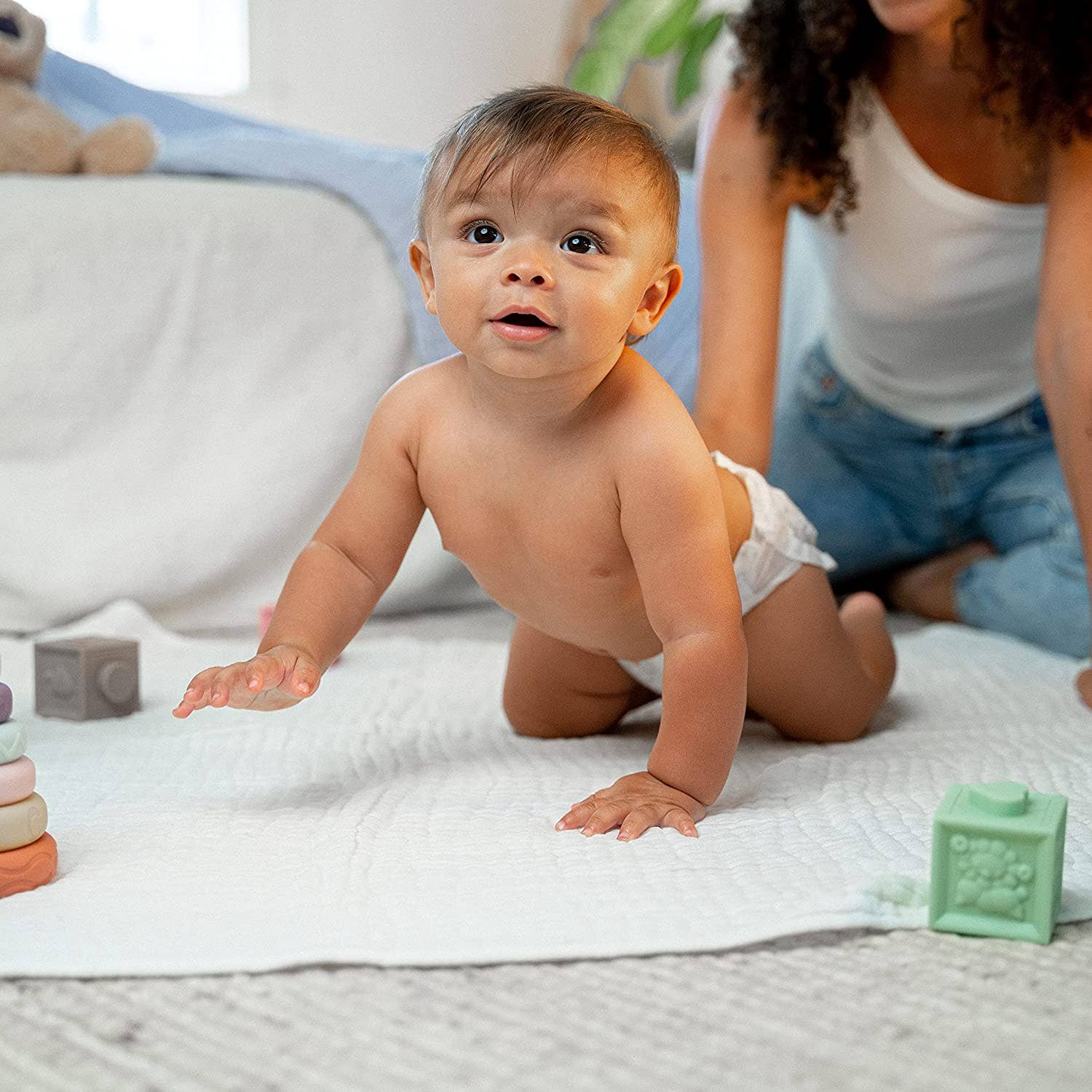 Happy baby crawling on a soft white muslin blanket, surrounded by colorful toys and a caring parent.