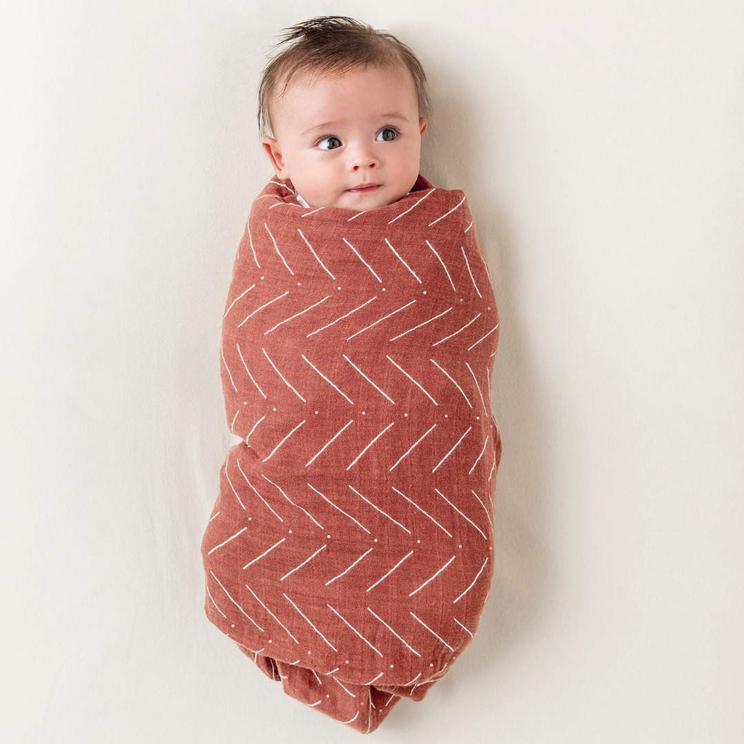 A happy baby wrapped in a soft, rust-colored swaddle with a white zigzag pattern, showcasing comfort and style.