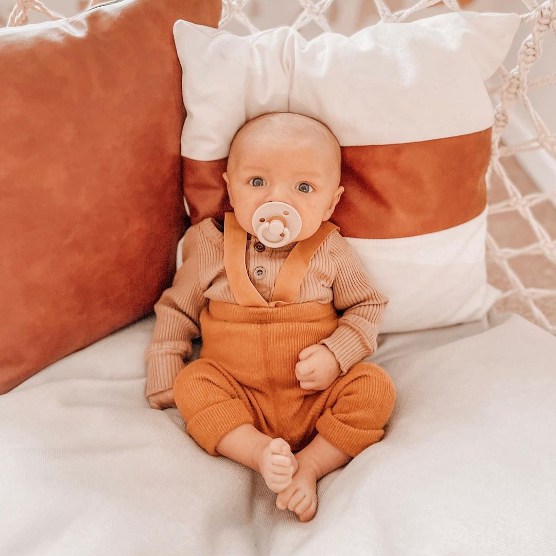 Baby relaxing on cushions with Itzy Ritzy natural rubber pacifier in coconut color, showcasing stylish baby fashion.