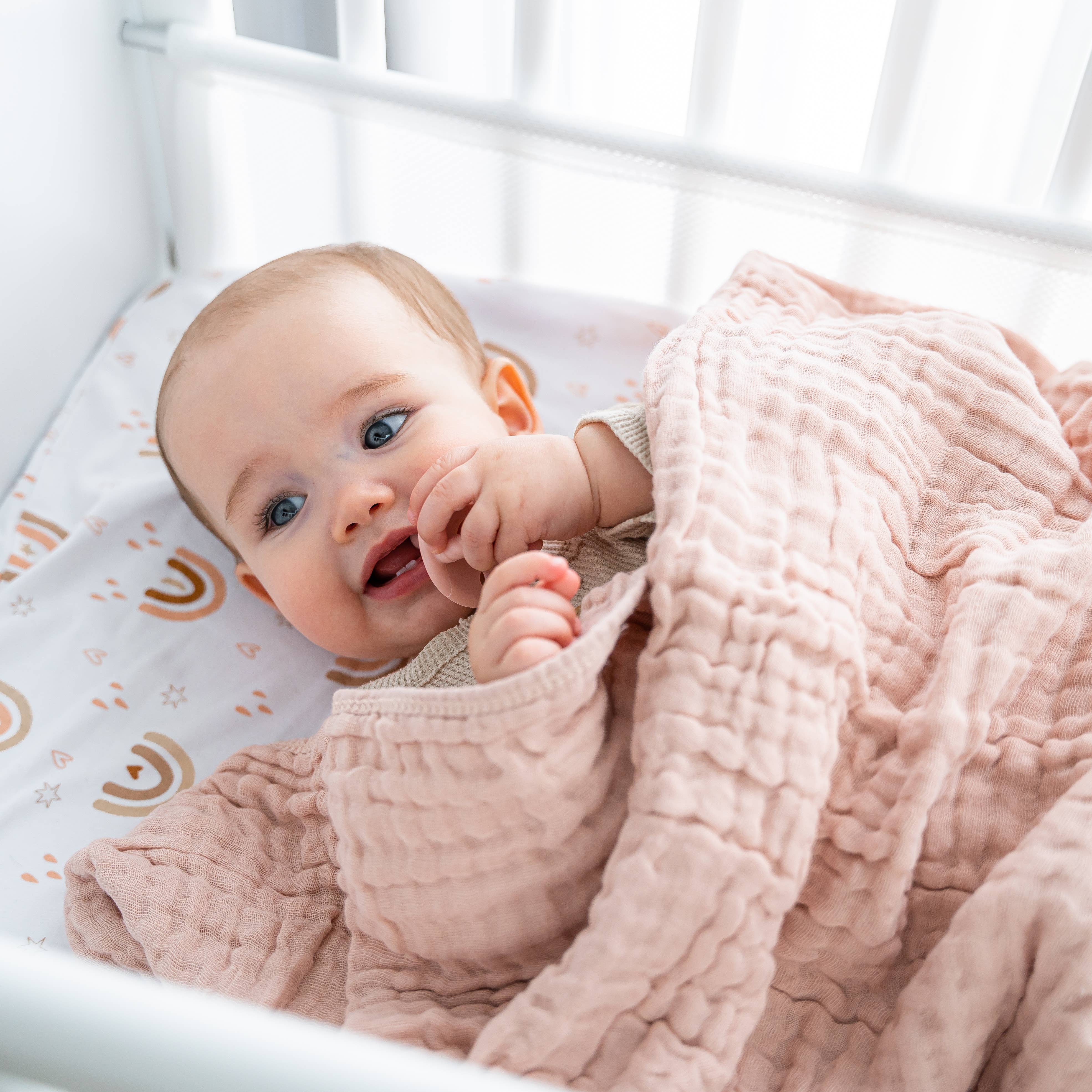 Smiling baby wrapped in a soft pink muslin cotton blanket in a crib, showcasing comfort and warmth.