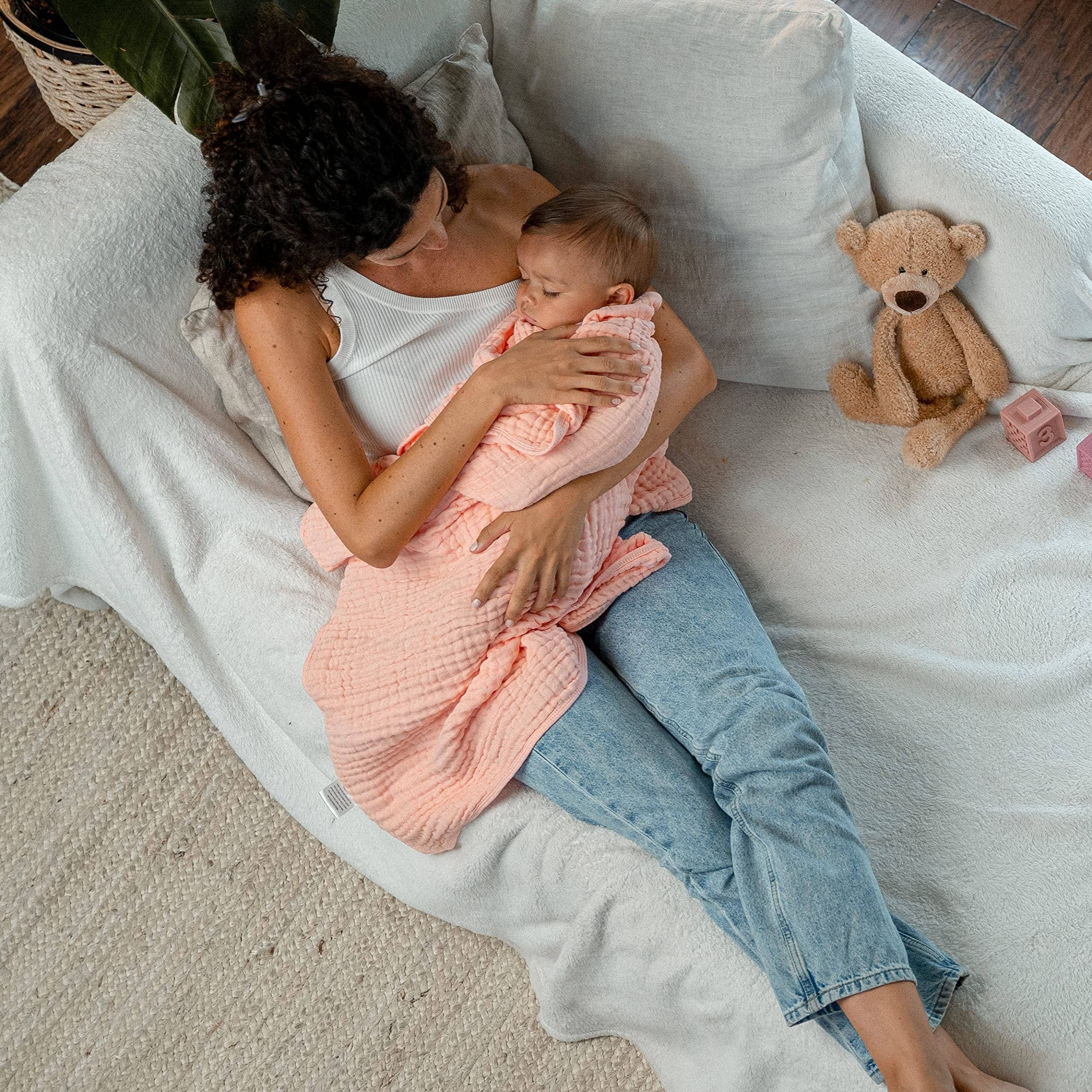 Mom cuddles baby in cozy pink muslin blanket on a soft couch, with a teddy bear nearby, creating a nurturing atmosphere.
