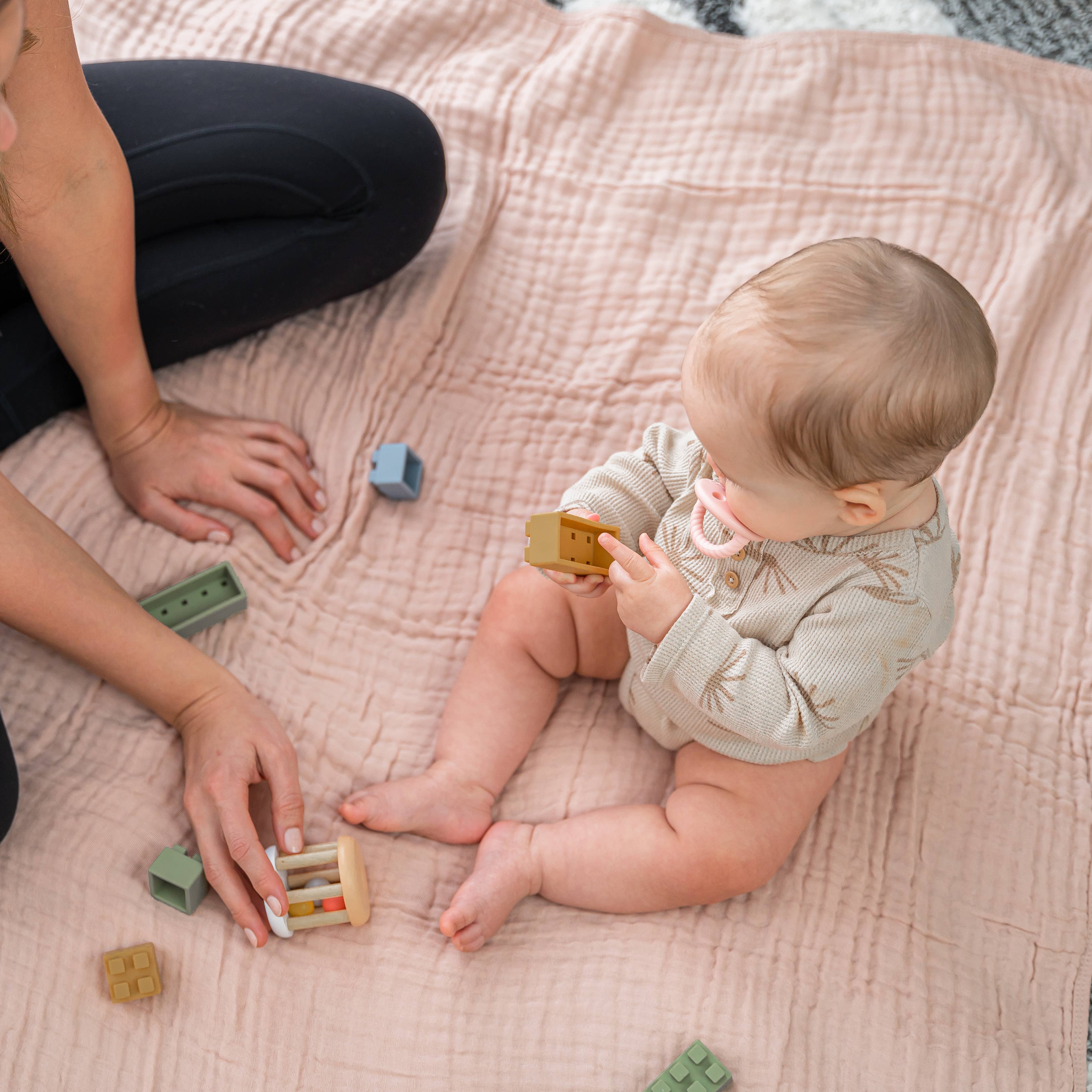 A baby sitting on a soft muslin blanket playing with colorful wooden blocks, enjoying quality playtime.