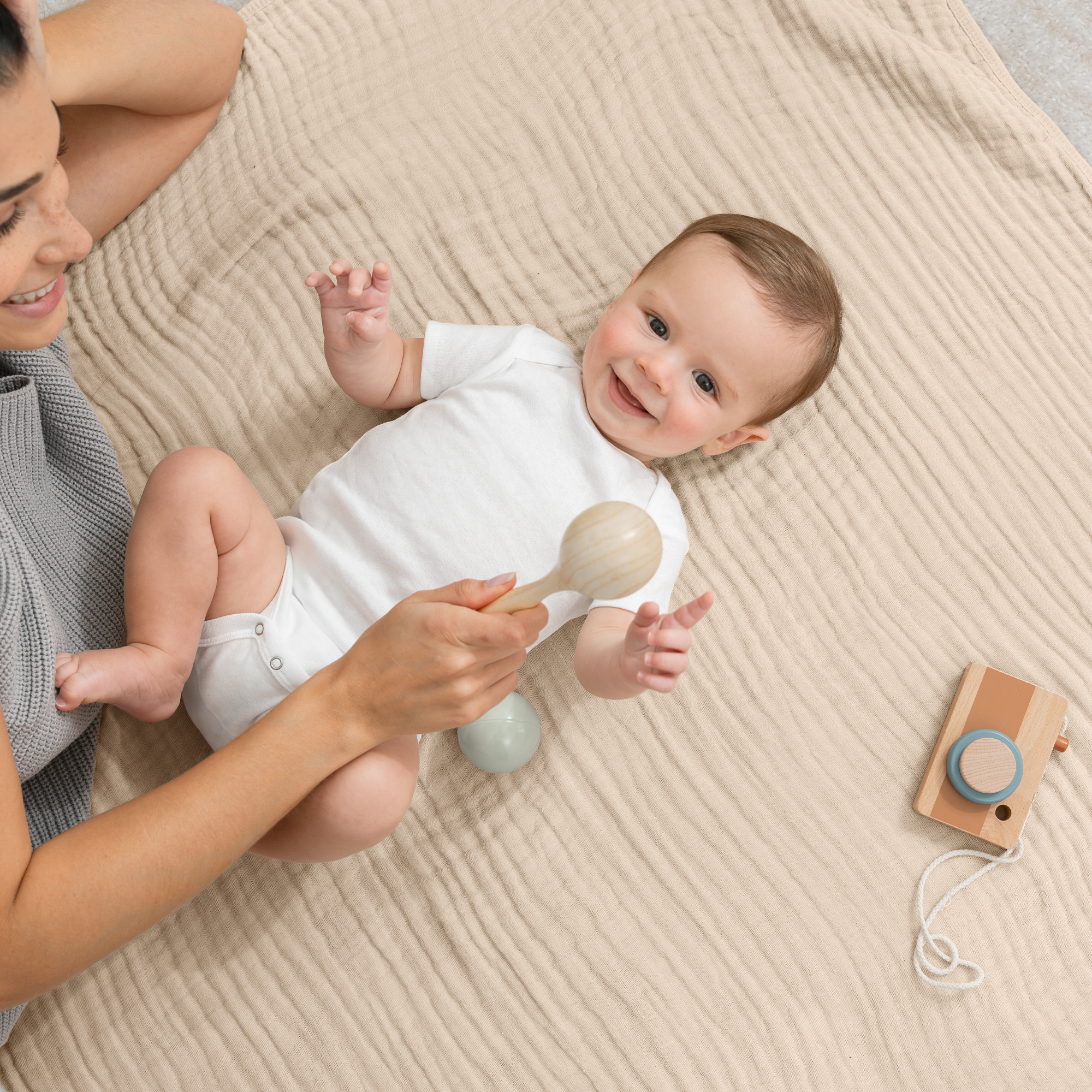 Happy baby playing with a wooden rattle on a cozy muslin cotton blanket with mom nearby.